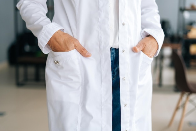 Foto recortada de close-up de médica irreconhecível em uniforme branco, segurando as mãos nos bolsos em pé no escritório do hospital. Mulher praticante posando no local de trabalho. Conceito de trabalho médico.