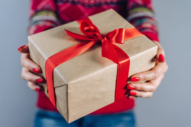 Foto recortada de garota em um suéter de malha com padrão tradicional de natal segurando uma caixa de presente embrulhada em papel artesanal e decorada com fita de cetim vermelha