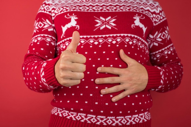 Foto recortada de homem de suéter de natal vermelho e branco segurando seu estômago e fazendo o polegar para cima em fundo vermelho isolado