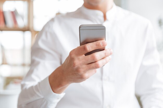 Foto recortada de jovem bonito vestido com uma camisa branca, usando telefone celular. Coworking.