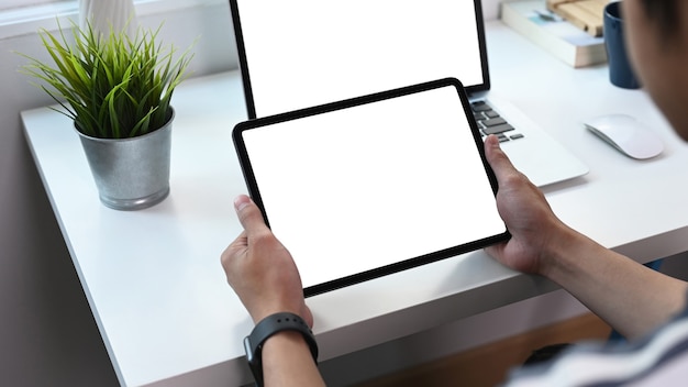 Foto recortada de jovem segurando o tablet digital com tela branca e usando o computador portátil na sala de escritório em casa.