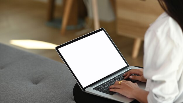 Foto recortada de mãos de freelancer de mulher digitando no laptop com tela branca enquanto está sentado no sofá.