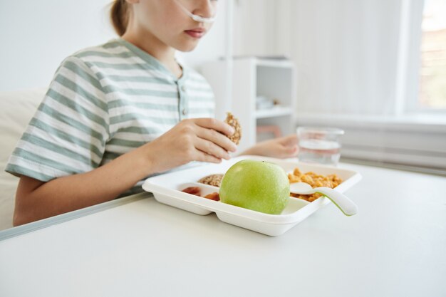 Foto recortada de menina comendo um almoço saudável em um quarto branco de hospital, plano de fundo com espaço de cópia