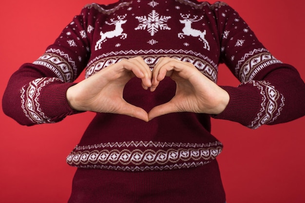 Foto recortada de menina de suéter de natal vermelho e branco fazendo coração com dedos em fundo vermelho isolado