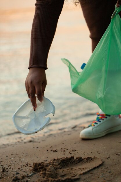 Foto recortada de mulher africana coletando lixo derramado de areia na praia em saco plástico verde Poluição ecológica