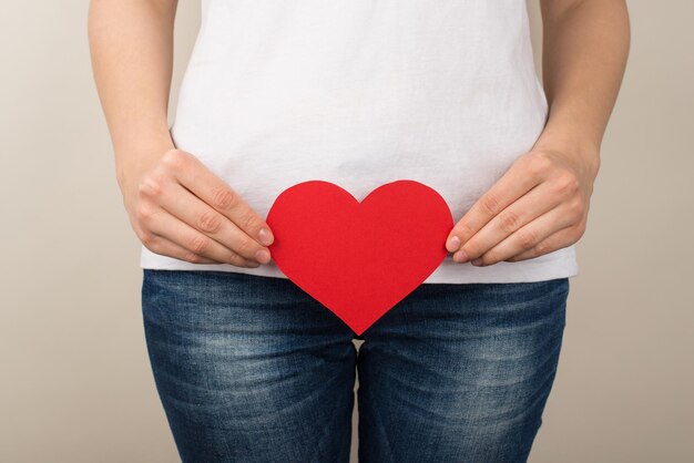 Foto recortada de mulher de camiseta branca e jeans segurando coração de papel vermelho perto da virilha em fundo cinza isolado
