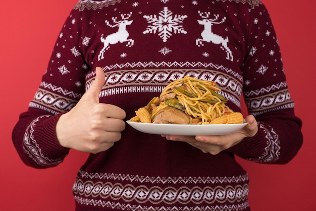 Foto recortada de mulher de suéter de natal vermelho e branco segurando um prato grande de comida e fazendo o polegar para cima em fundo vermelho isolado