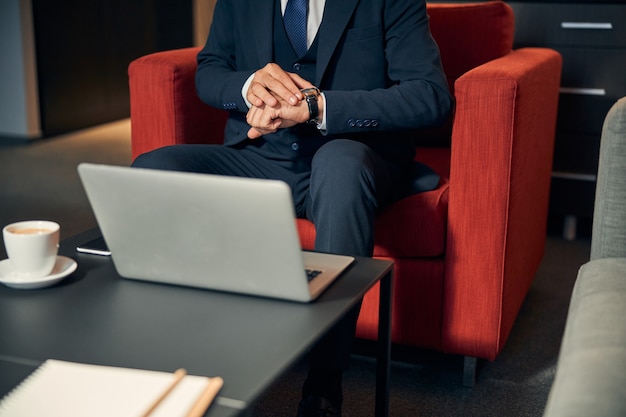 Foto recortada de um empresário sentado na poltrona na zona lounge e tocando o relógio no pulso. Xícara de café e um laptop na mesa