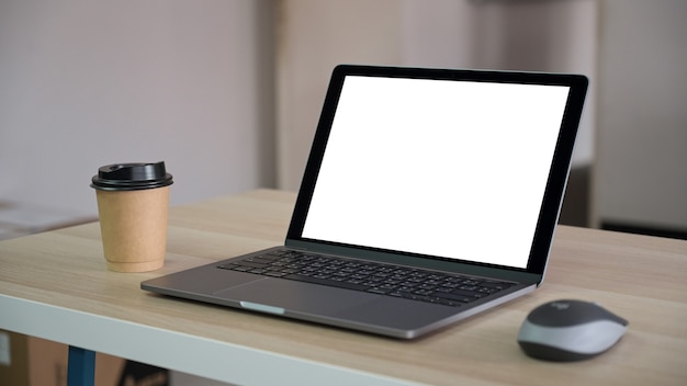 Foto recortada de um laptop com tela em branco e uma caneca de café marrom para viagem na mesa do escritório