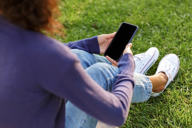 Foto recortada de uma jovem africana sentada ao ar livre no parque usando telefone celular.