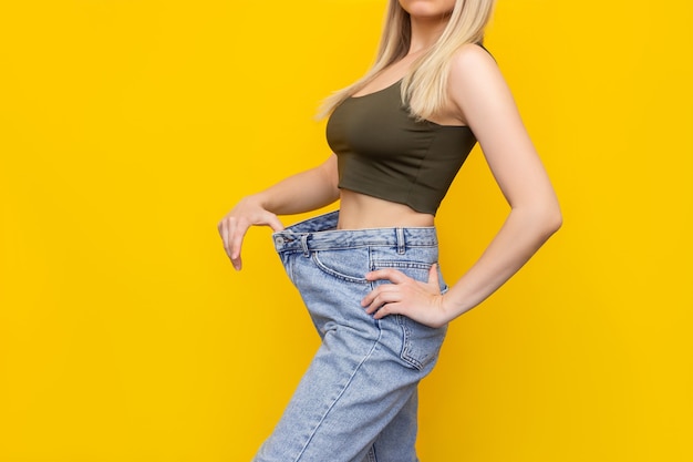 Foto recortada de uma jovem loira esguia, branca, vestida com jeans largos e top curto, demonstra perda de peso bem-sucedida isolada em uma parede de cor amarela