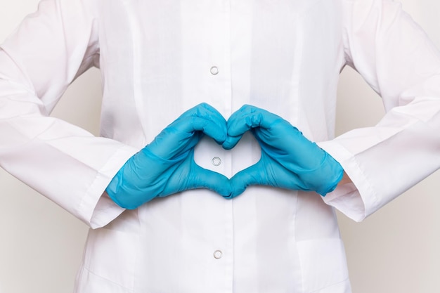 Foto recortada de uma médica de jaleco branco formando uma forma de coração com as mãos em luvas azuis