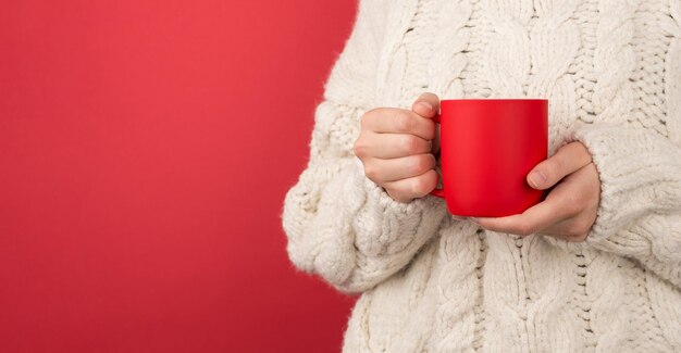 Foto recortada de uma mulher de suéter branco segurando uma xícara vermelha de bebida quente em fundo vermelho isolado com espaço para cópia