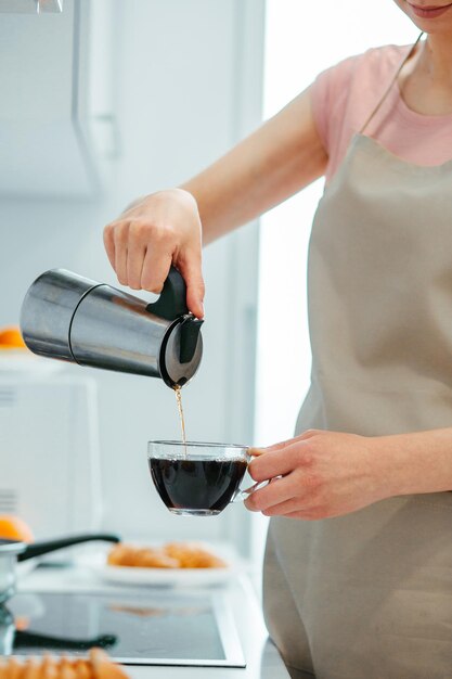 Foto recortada de uma mulher sorridente derramando cuidadosamente café de uma panela em pé na cozinha