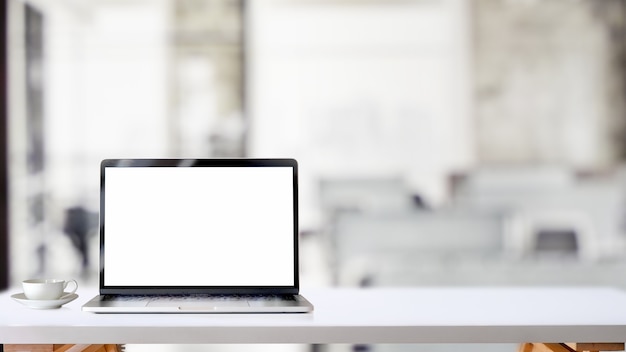 Foto recortada do espaço de trabalho simples com laptop de tela em branco e xícara de café na mesa branca com fundo de sala de escritório turva