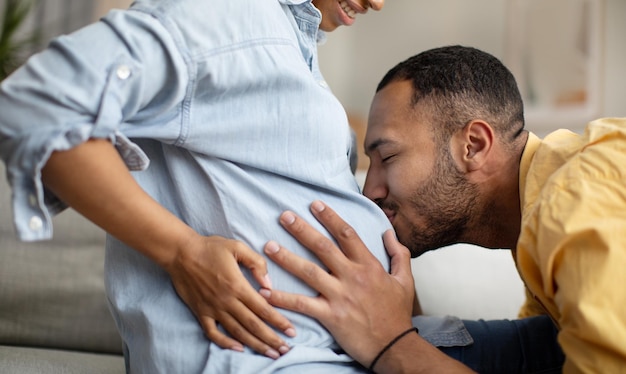 Foto recortada do marido afro-americano beijando a barriga da esposa no interior