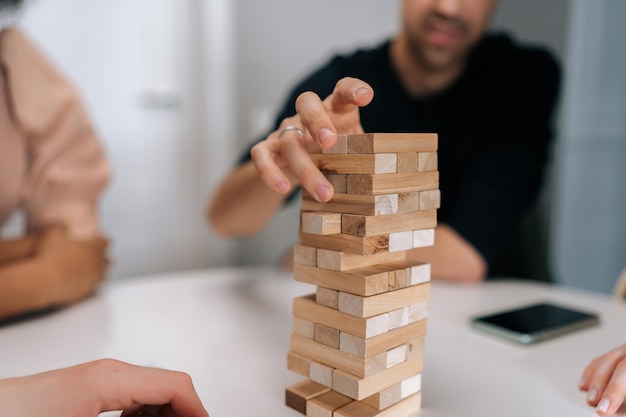 Foto recortada em close de um homem irreconhecível removendo cuidadosamente o bloco de madeira da torre instável empilhada durante a brincadeira com os amigos