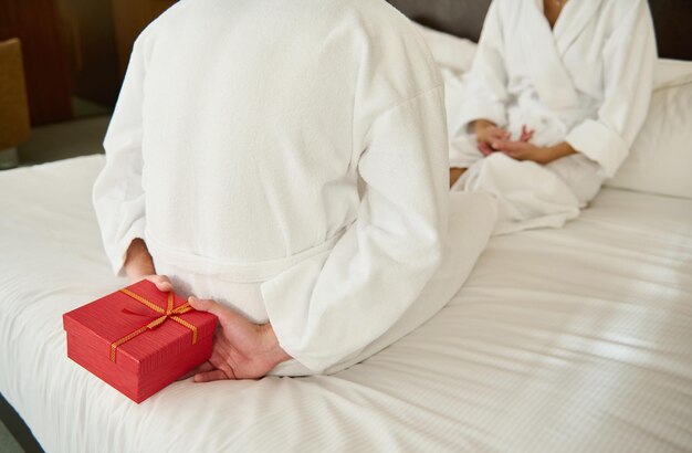 Foto recortada de un hombre irreconocible en bata de baño de felpa blanca sentado frente a su amante en la cama y escondiendo una caja de regalo roja, lo que la convierte en una sorpresa para el día de San Valentín. Los amantes dan regalos para las fiestas.