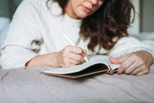 Foto recortada de una joven morena adulta de cuarenta años de talla grande con cuerpo positivo en suéter de punto blanco escribe notas en el planificador en la cama en el hogar