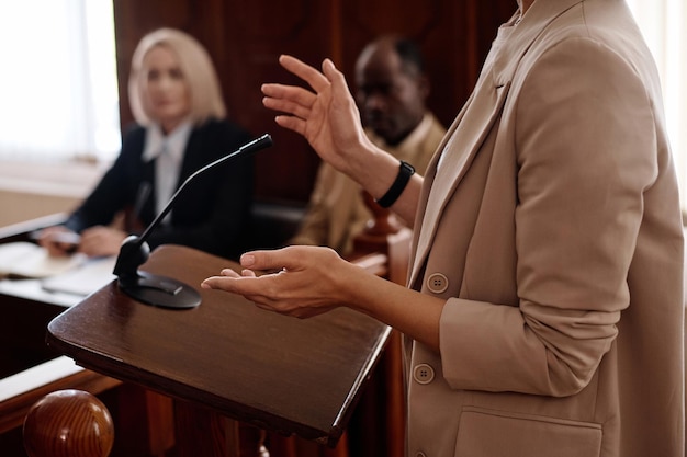 Foto recortada de una joven testigo femenina con chaqueta beige de pie junto a la tribuna con un micrófono