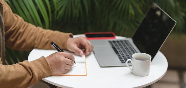 Foto recortada joven trabajando en su proyecto con computadora portátil y suministros de oficina en mesa blanca