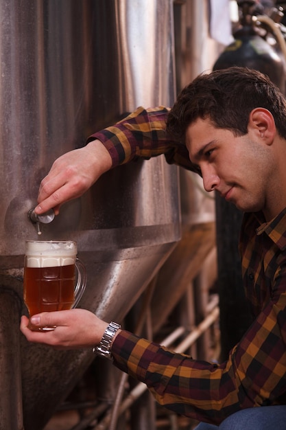 Foto recortada vertical de um cervejeiro profissional derramando cerveja recém-fabricada do tanque de metal em uma caneca