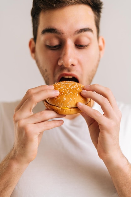 Foto recortada verticalmente de um jovem faminto gostando de comer um delicioso hambúrguer em fundo branco isolado