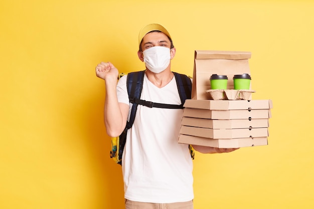 Foto de un repartidor con gorra amarilla, camiseta blanca y mascarilla protectora sosteniendo pizza y café con los puños apretados regocijándose posando aislado en el fondo amarillo