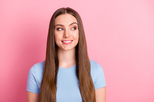 Foto retrato de uma garota curiosa com longos cabelos castanhos sorrindo, olhando para o espaço em branco isolado em um fundo de cor rosa pastel.