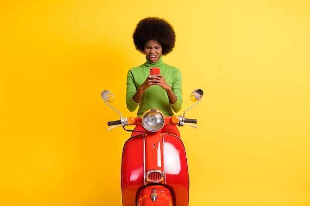 Foto retrato de uma jovem morena afro-americana com roupa casual em uma motocicleta vermelha segurando um celular, olhando com nojo para uma mensagem sms isolada em um fundo colorido amarelo vívido