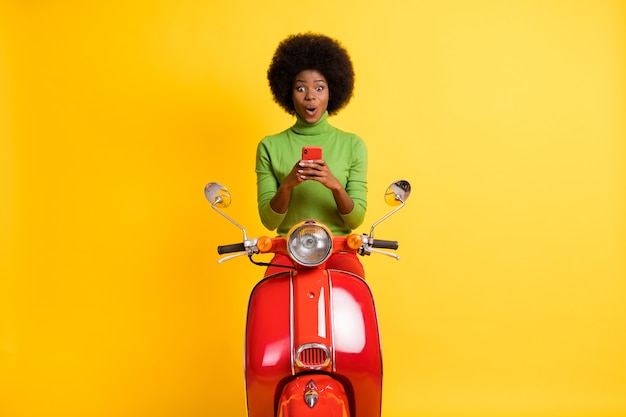 Foto retrato de joven morena sorprendida mujer afroamericana emocionada en bicicleta roja sosteniendo teléfono celular aislado sobre fondo de color amarillo vivo