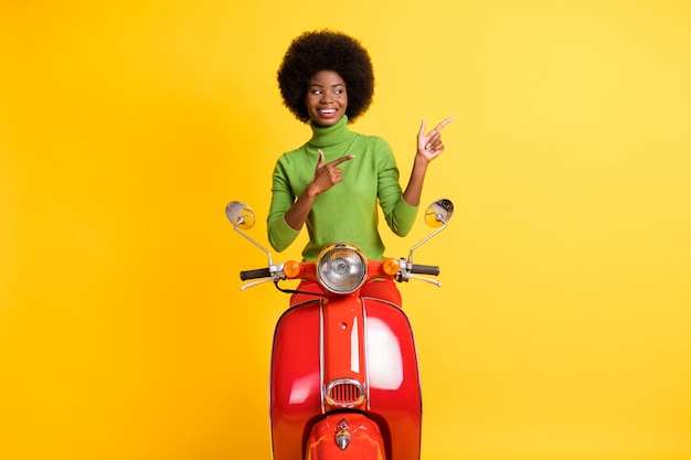 Foto retrato de joven motociclista de piel negra conduciendo apuntando con dos dedos hacia el lado aislado sobre fondo de color amarillo vivo