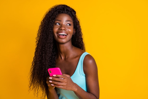 Foto retrato de joven mujer afroamericana rizada sosteniendo el teléfono en dos manos riendo vistiendo camiseta azul aislado sobre fondo de color amarillo brillante