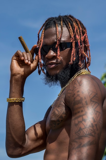 Foto de retrato de modelo masculino africano con tatuajes rastas cadenas de oro y cigarro en las manos