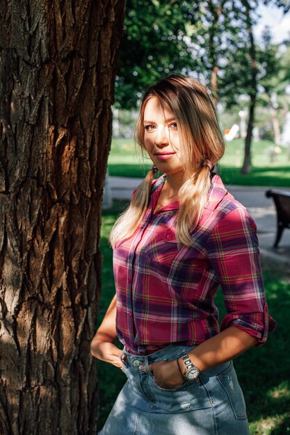 Foto de retrato de mujer rubia brillante adulta de pie en el parque cerca de roble con las manos en los bolsillos vistiendo ...