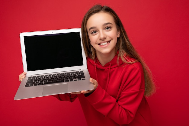 Foto de retrato de primer plano de hermosa niña sonriente feliz con cabello largo vistiendo sudadera roja con capucha sosteniendo computadora portátil