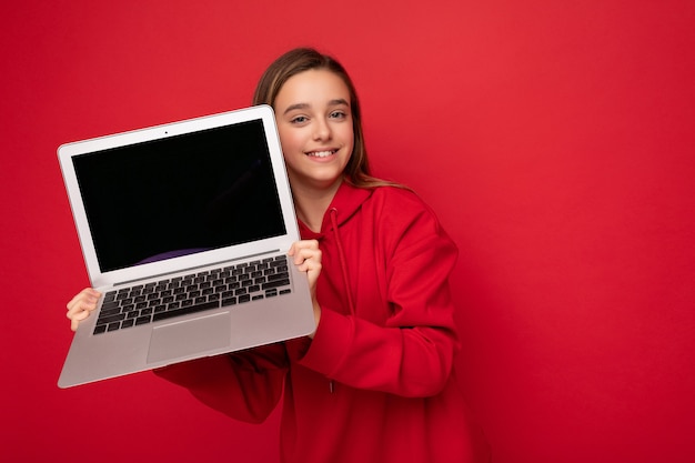 Foto de retrato de primer plano de hermosa niña sonriente feliz con cabello largo vistiendo sudadera roja con capucha sosteniendo computadora portátil