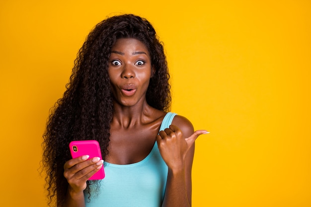 Foto retrato sorprendido sorprendido rizado mujer afroamericana afroamericana que señala con el dedo pulgar copyspace sosteniendo el teléfono en la mano vistiendo casual azul camiseta sin mangas aislado fondo de color amarillo brillante