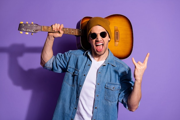 Foto de rockero atractivo alegre gritando de moda que le muestra el signo de rock dedos con cuernos sosteniendo la guitarra acústica en el hombro con gafas de mezclilla aisladas de fondo de color púrpura vivo