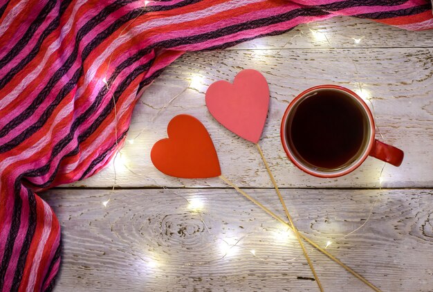 Foto romântica com uma caneca de chá, um lenço listrado, dois corações vermelhos e luzes mágicas em um fundo de madeira