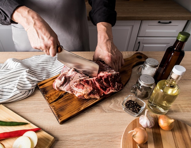 Una foto sin rostro de un hombre con un delantal gris con un hacha corta un gran trozo de carne en los huesos el concepto de cocinar carne en la cocina de casa