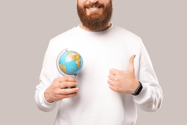 Foto sem rosto de um homem mostrando o polegar para cima e segurando um pequeno globo terrestre Studio atirou sobre fundo cinza