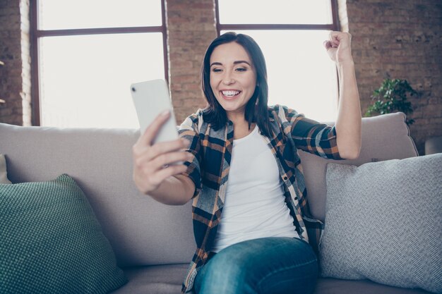 Foto de señora bonita sosteniendo el teléfono leyendo las mejores noticias sobre el proyecto de inicio levantando el puño sentado en un sofá acogedor con ropa informal apartamento en el interior