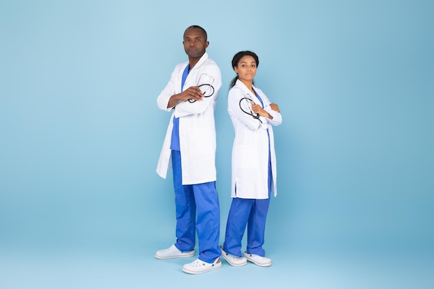 Foto de serios profesionales médicos negros masculinos y femeninos posando con los brazos cruzados usando batas de laboratorio de cuerpo entero