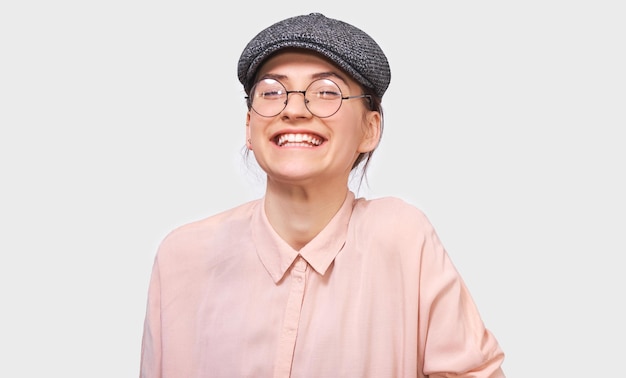 Una foto sincera de una joven feliz que sonríe ampliamente viste una camisa rosa y gafas transparentes redondas Una estudiante bonita se siente alegre y posa sobre una pared blanca del estudio Concepto de personas y emociones