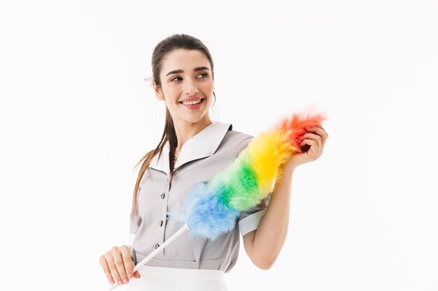 Foto de sirvienta sonriente de 20 años vestida con uniforme sosteniendo un plumero de colores mientras hace las tareas del hogar y la limpieza de la habitación aislada sobre la pared blanca