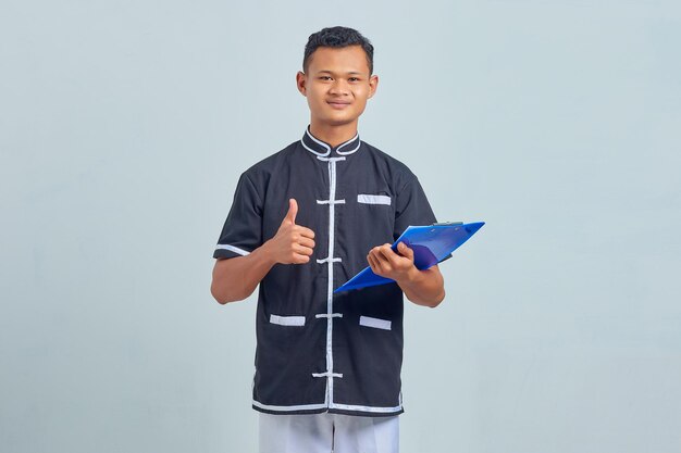 Foto de sonriente joven asiático vistiendo uniforme de karate sosteniendo el portapapeles mostrando el pulgar hacia arriba