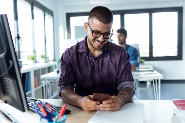 Foto de sonriente joven empresario con su teléfono inteligente mientras trabaja con la computadora en la oficina de inicio moderna.