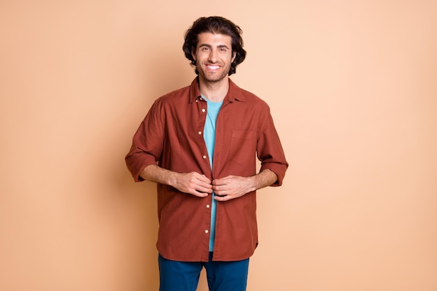 Foto de sonriente joven lindo desgaste casual traje marrón abroche el botón de la camisa aislado de fondo de color beige