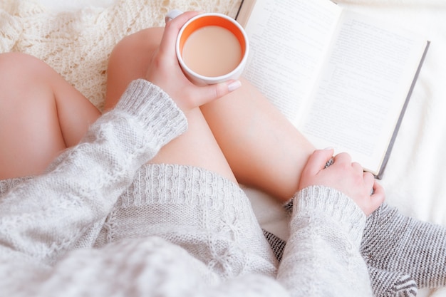 Foto suave de una mujer con un acogedor suéter en la cama con el libro antiguo y una taza de té con leche en sus manos